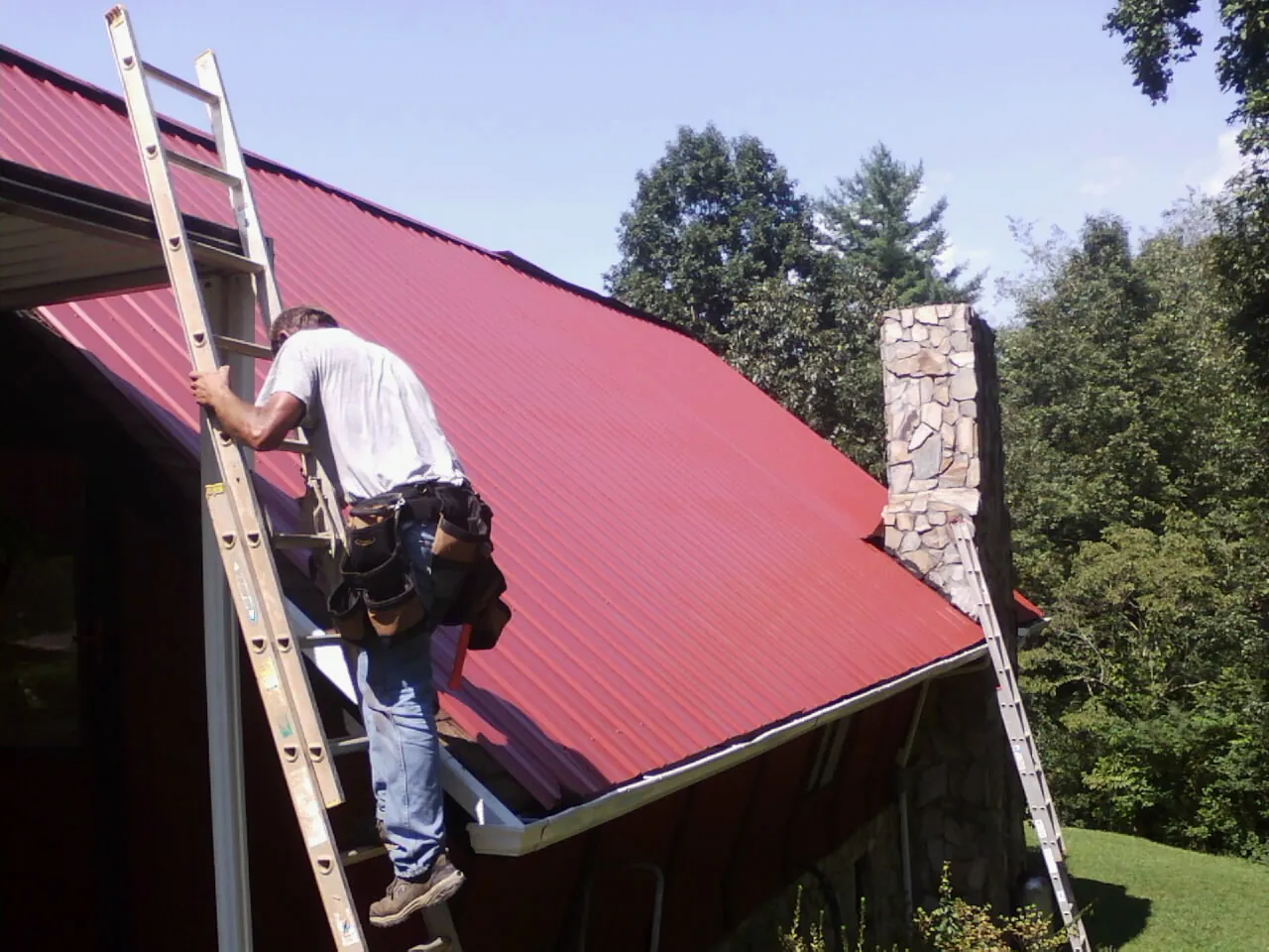 Roofer on a ladder beside a red metal roof and stone chimney — Soffit & Fascia Repair by Roofing Brothers in Goodland